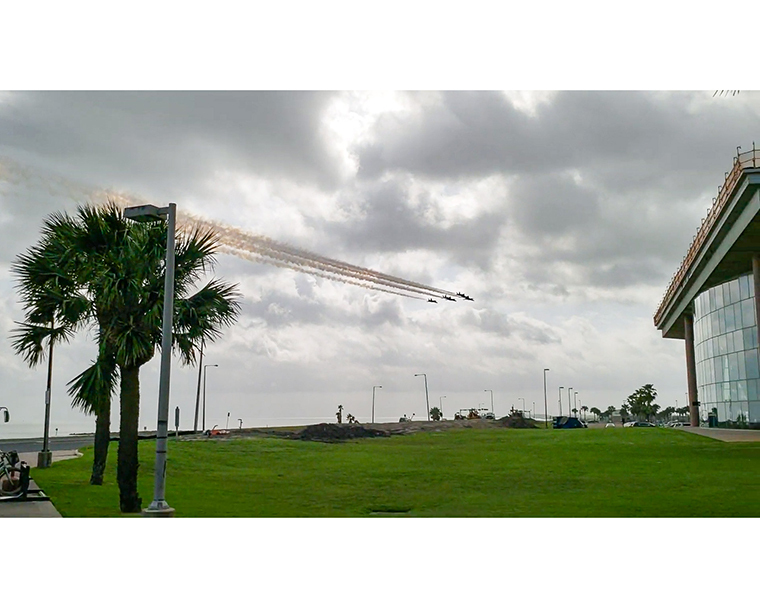 Color photograph of a group of small airplanes flying low over the shoreline. The sky is overcast and the group of planes leave a trail of smoke. The foreground includes a grassy lawn, palm trees, and buildings.