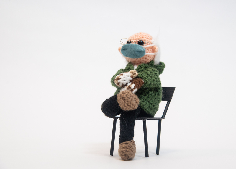 Crocheted man bundled in warm clothing including mittens seated in a chair.