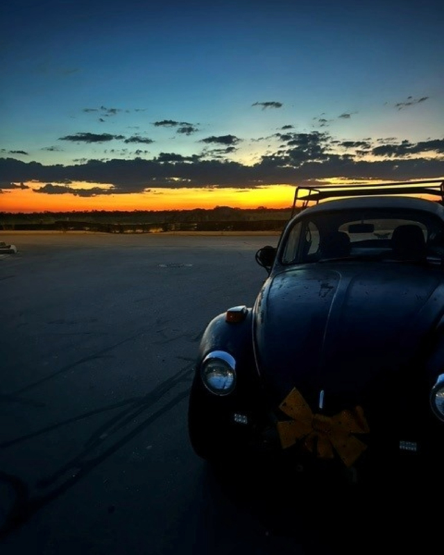 A vintage black car with a yellow bow sits in the foreground against a colorful sunset with blue and orange hues in the sky.