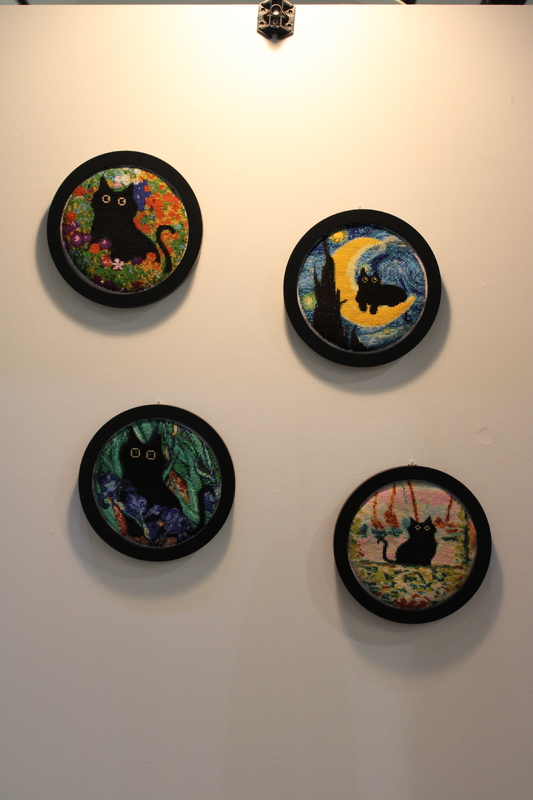 Four embroidery hoops with black cat cross-stitch art in varied colorful backgrounds mounted on a wall.
