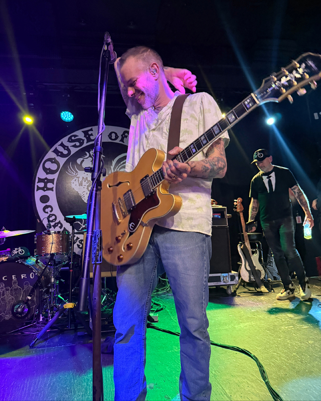 A guitarist in a patterned shirt plays on stage with colorful lighting, while a band member stands behind him. A drum set is visible in the background.