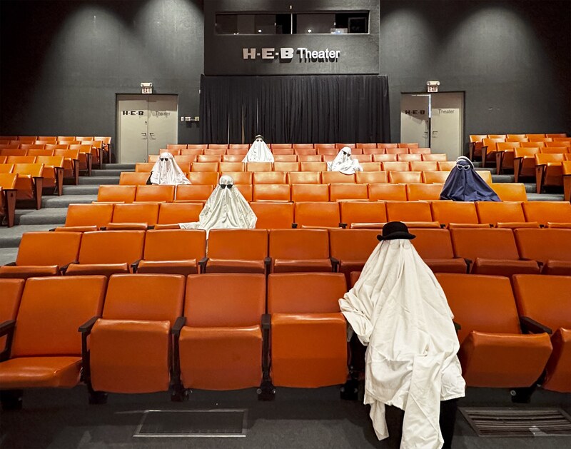 Artist: Alexis De León. A 12 by 15-inch archival pigment print, titled Sticking Around for the Stinger. The print captures the inside at a Theater with orange stadium seating. Six figures are scattered throughout the theater draped in fabric. Five with light fabric, one with dark covering. All wear hats and glasses on top of the draped covering. The perspective is from the front of the theater, looking at the audience.
