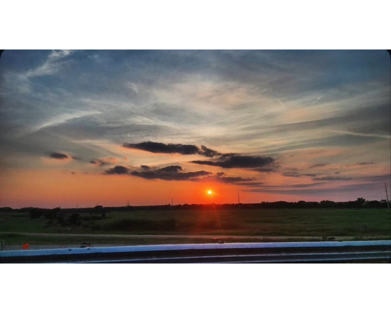 A sunset over a rural landscape with an orange sun near the horizon and pastel-colored clouds.