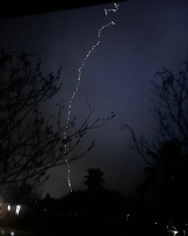 A jagged lightning bolt strikes downward in a dark, stormy sky, with bare tree branches silhouetted in the foreground.