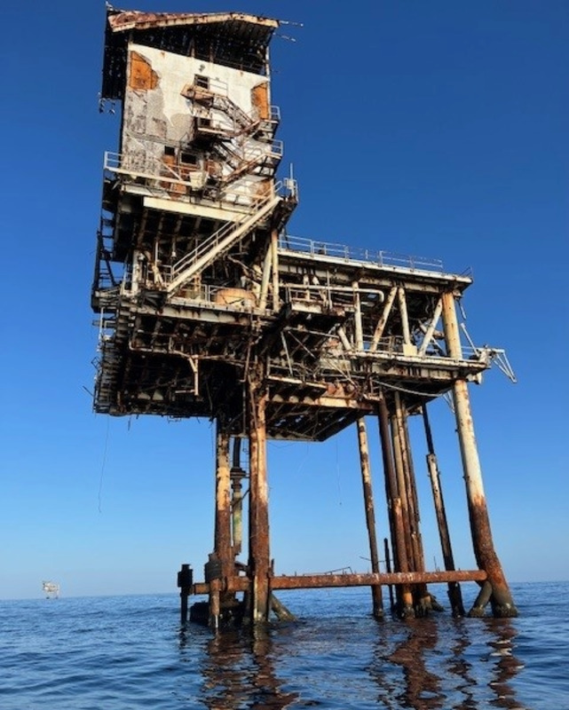 A large, several storied structure is elevated on metal pilings above a body of water. The structure includes, stairways, handrails, and piping. The surface of the building and the supporting pilings are showing signs of rust and damage. A similar structure can be seen in the far distance. The two structures are surrounded by water and a clear blue sky.