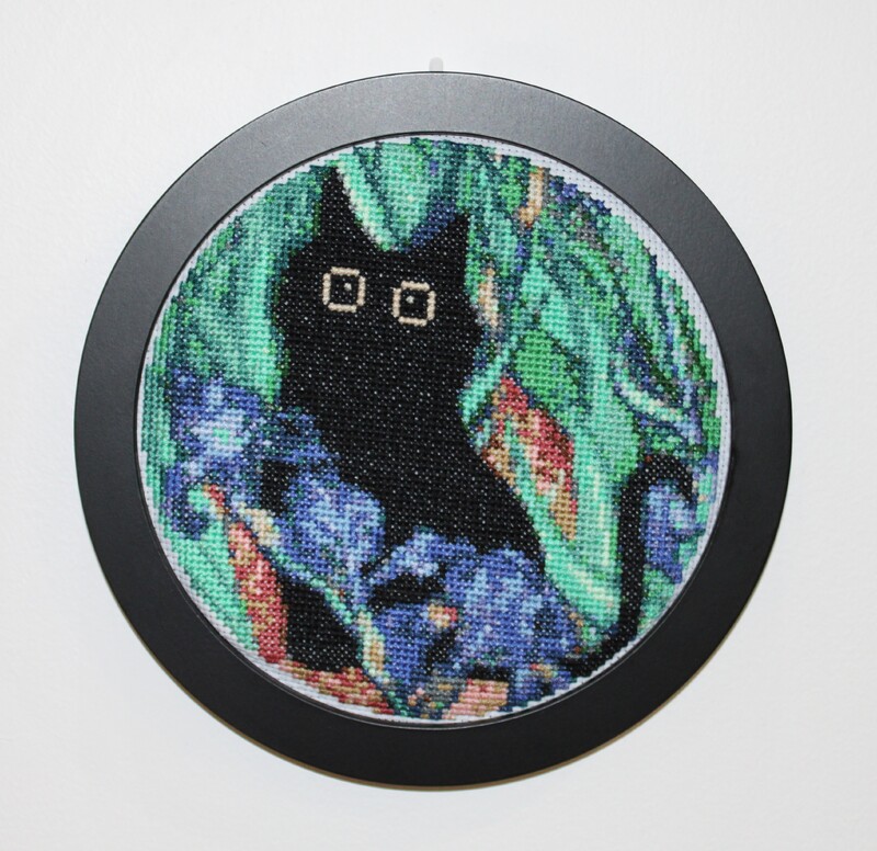 Embroidery hoop with black cat cross-stitch art in a green leafy background near blue irises.
