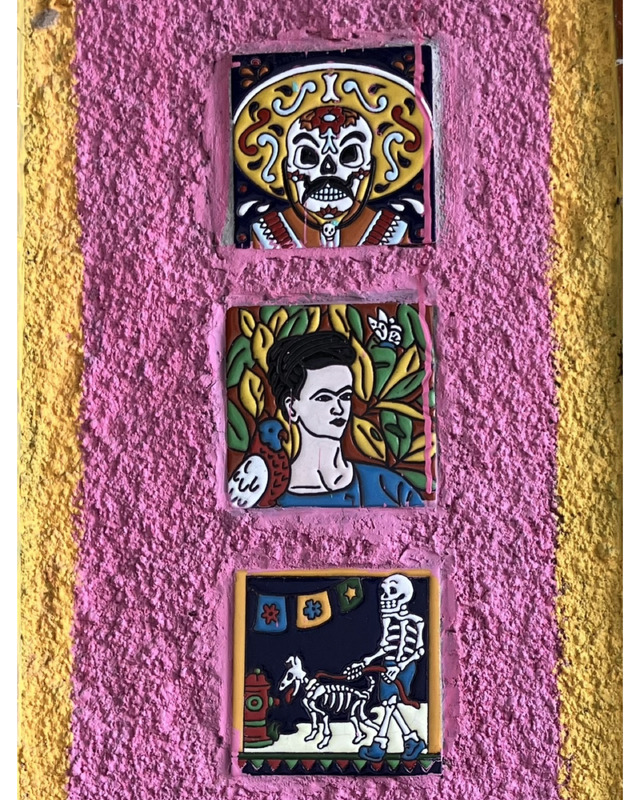 Three colorful ceramic tiles on a pink wall: a decorative skull, a portrait of a woman reminiscent of Frida Kahlo, and a skeleton walking a dog.