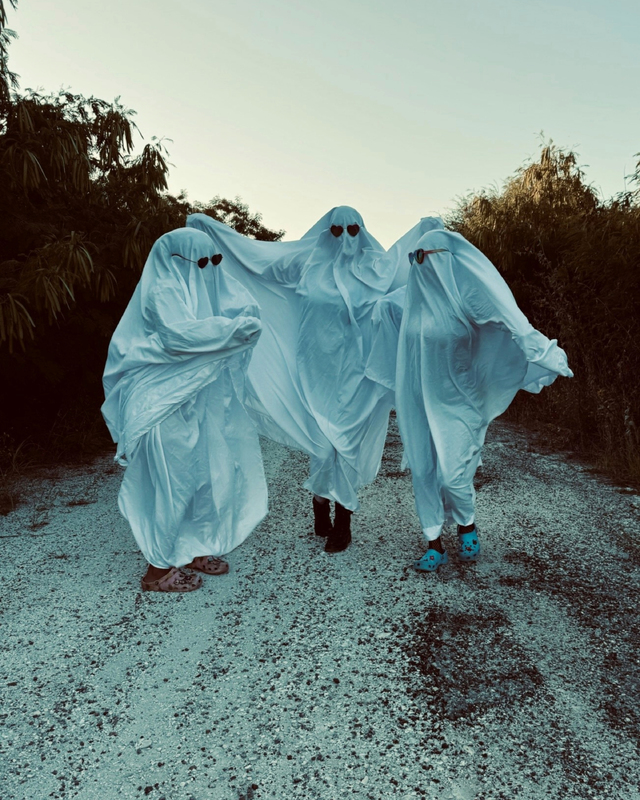 Three people covered in white sheets with sunglasses pose as ghosts on a gravel path surrounded by trees, wearing casual shoes.
