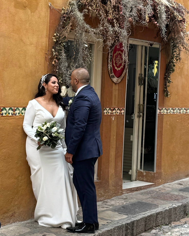 A smiling figure in a long white gown, with delicate white headpiece. They are holdings a floral bouquet, leaning against a brown building. Another figure stands in facing the first, dressed in a dark blue suit, with a white floral boutonniere in their left pocket. The stone sidewalk and street are visible. The couple are close to a glass doorway and window with dried foliage in a garland above the openings. A red round symbol decorates the wall and a decorative tile trim runs the length of the outside of the building.