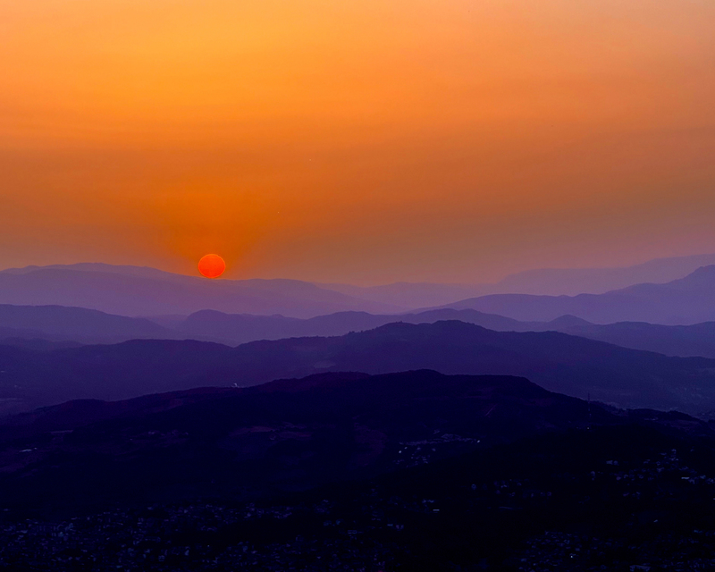 A vibrant red sun sets over layered purple and orange mountains, creating a tranquil gradient sky.