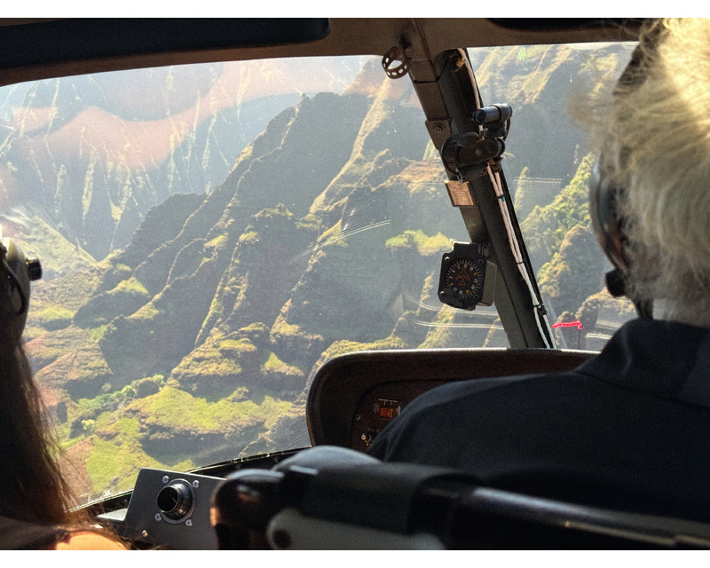Interior view of a helicopter cockpit with two people wearing headsets, overlooking steep green mountains below.