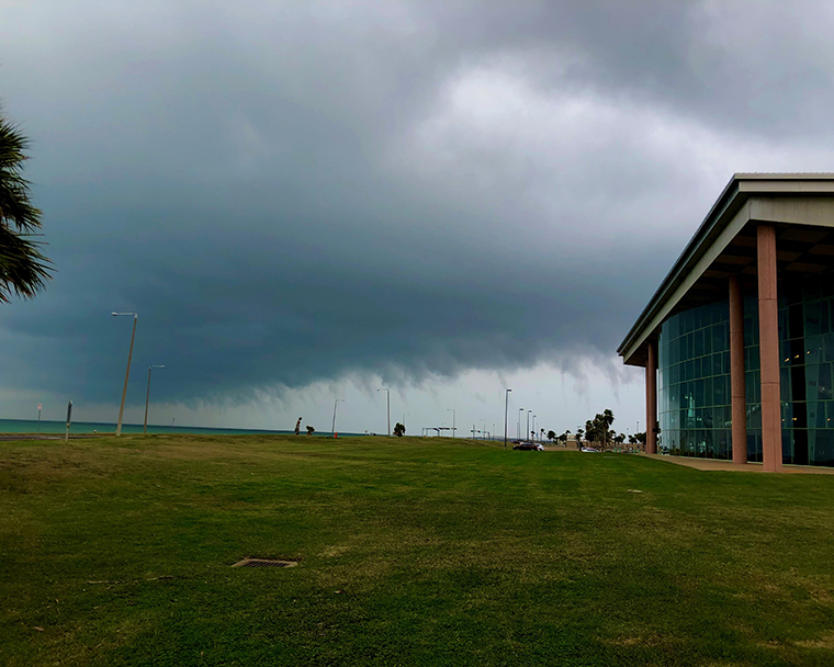 Color photograph of low wispy storm clouds. The landscape includes an expanse of grass, a building, and a bay.