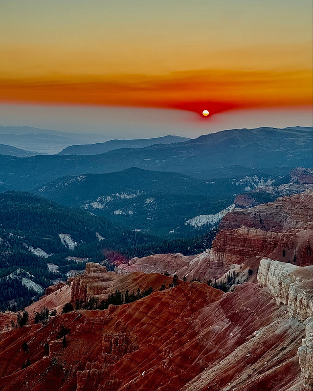 Sunset over a mountainous landscape with layers of red and brown hills, dotted with green trees, beneath an orange and blue sky.