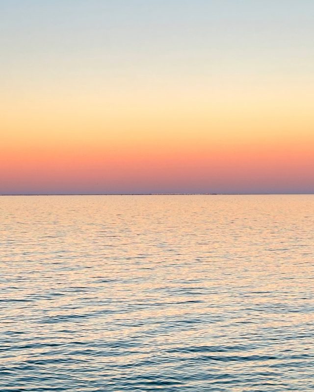 A calm seascape at twilight, displaying a smooth water surface reflecting soft hues of pink and blue, with a clear horizon.