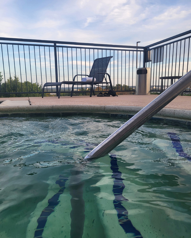 Close view of a hot tub with clear water, a black lounge chair with a towel, and a pair of flip-flops nearby, set against a distant landscape and cloudy sky.