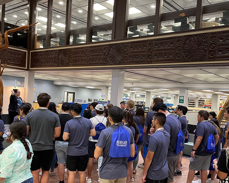 Color photograph of a group of people inside a building facing a presenter. The photograph includes architectural details of the space. 