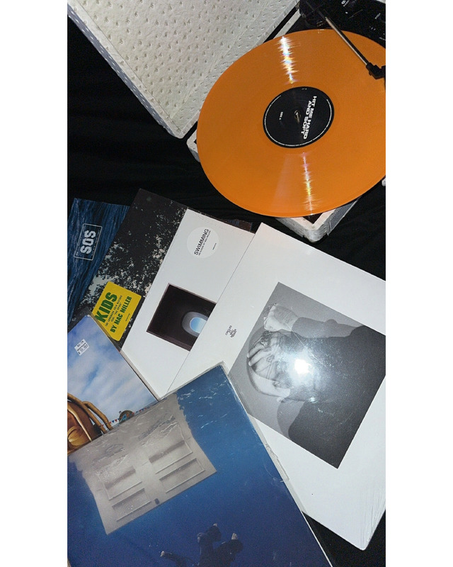A collection of vinyl records on a dark surface, with an orange record on a turntable, surrounded by various album covers including "Swimming" and a yellow sticker saying "KIDS BY MAC MILLER."