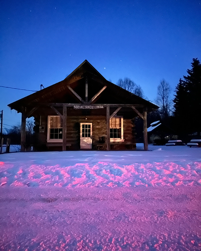 A rustic log building labeled "Social Hall 1902" glows at twilight, surrounded by snow reflecting pink and purple light.