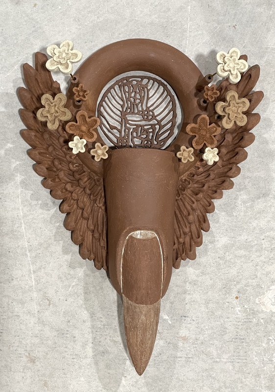 Artist: Leandra Urrutia. 13 by 7 by 3-inch ceramic and wire sculpture. The sculpted piece includes a fingertip with sharpened nail flanked by wings and flowers. The sculpture has a circular top with a delicate latticed structure in the center.