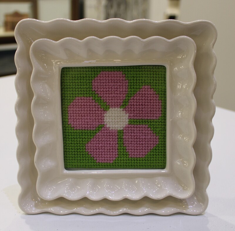 Cross-stitched pink flower with white center on a green background, framed in a wavy-edged cream ceramic frame.