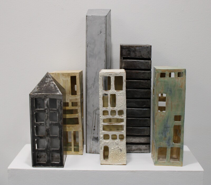 A group of ceramic sculptures resembling a city skyline on a white shelf, featuring simplified skyscraper forms in various colors and textures.

