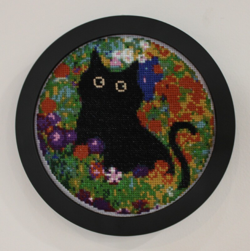 Embroidery hoop with black cat cross-stitch art in colorful floral background.
