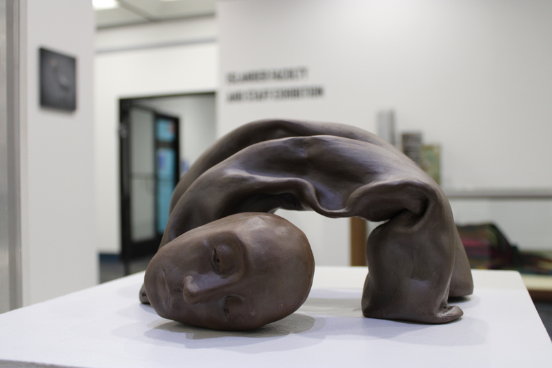 The image shows a sculpture placed on a white pedestal inside a gallery space. The sculpture is crafted from a material resembling bronze or dark clay, depicting a human figure in an arched position.
