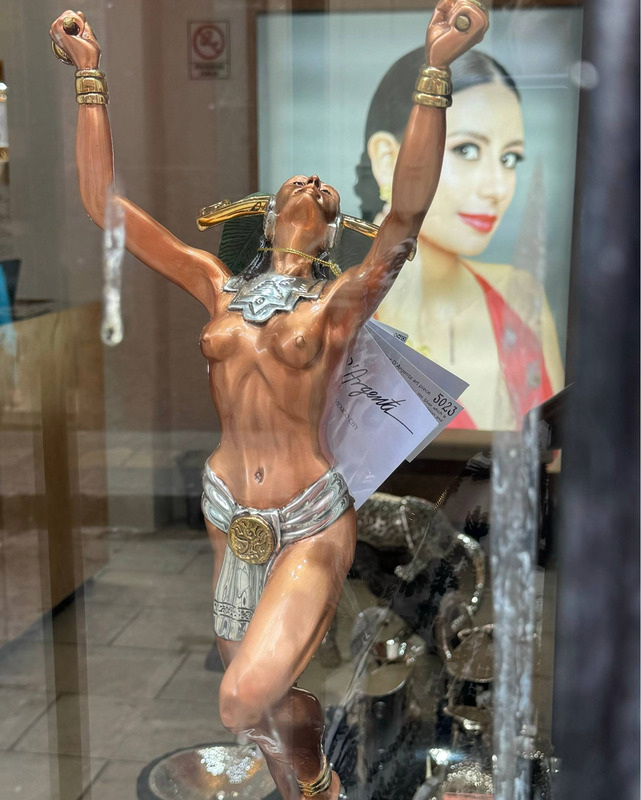 A bronze colored sculpture of a female figure with arms upraised. The figure is holding items in their hands and is adorned with bracelets, headpiece, and a large silver colored neck/throat plate. The figure wears a silver colored loincloth that drapes beneath a gold circular emblem in the center. One leg is raised at the knee and a tag can be seen hanging from a thread around the sculpture's neck. In the background, a large image of a smiling women in a red dress can be seen. The figure appears to be gazing at the viewer and is wearing lipstick that matches the dress. Her hair is slicked back and gathered at the neck. The entire image is seen through a slightly reflective surface - as if viewed through a window.