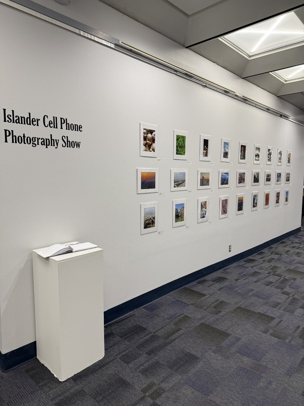 Interior view of a photographic exhibition titled "Islander Cell Phone Photography Show," featuring framed photographs on white walls.