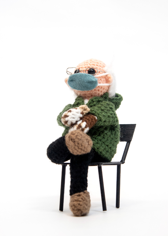 Crocheted man bundled in warm clothing including mittens seated in a chair. Closer view.