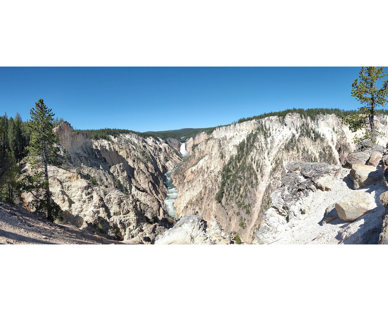 A distant waterfall cuts through tree topped rocky mountains. The falls are centered in the frame and extend down towards the viewer. The sides of the mountain and the foreground are bare rock. The image is bound a white border at the top and bottom of the frame.