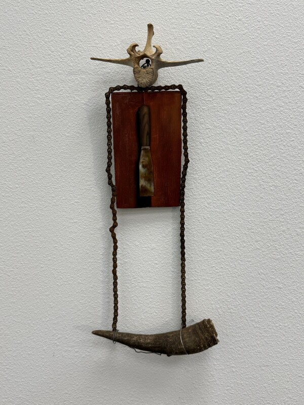 Artist: Joe Pena. 16 by 12-inch oil on panel and personal objects. A bone rests on top of a wooden painted panel. Hanging horizontally beneath is a horn, connected by wire. The piece is encircled with chain.