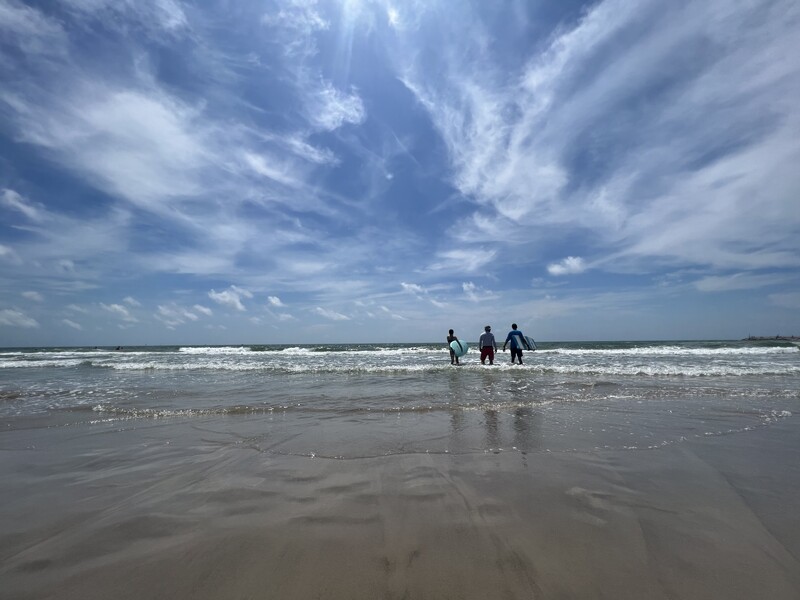 Three people with surfboards walking towards the ocean on a beach under a blue sky filled with wispy clouds.

