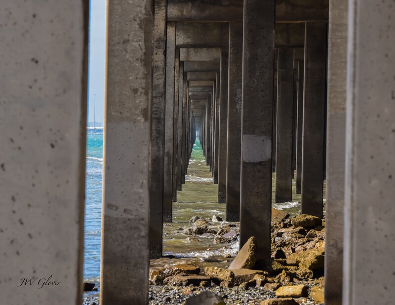Artist: Jesse Glover. 14 by 11-inch photograph printed on canvas. Signed photograph of a concrete structure over a shoreline. The perspective is from underneath the structure, looking in between the concrete pilings. A rocky shoreline gives way to waves and deeper water further in the frame.
