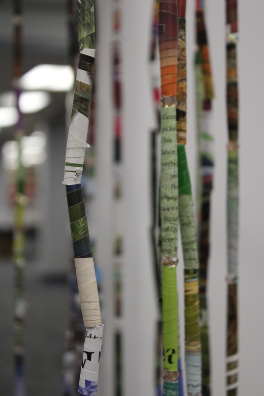 The installation consists of a circular frame from which numerous thin, colorful strips hang vertically. The strips appear to be made of tightly rolled paper, possibly from magazines or posters, and are suspended at various lengths, creating a curtain-like effect. T
