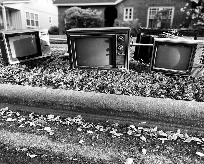 Three television sets and other equipment, resting on a lawn at the edge of a curbed street. Houses and shrubbery can be seen in the background. The image is in black and white and the image is lit from the right, casting shadows on the equipment. Tree leaves are scattered in the street.