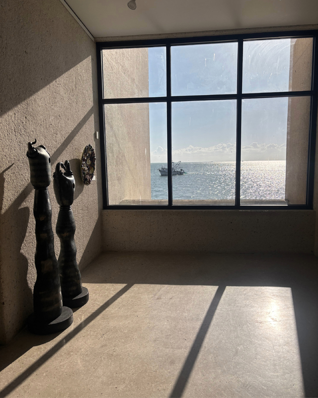 Two abstract sculptures positioned against a textured wall with a large window view of a fishing boat on the ocean, casting shadows on the floor.