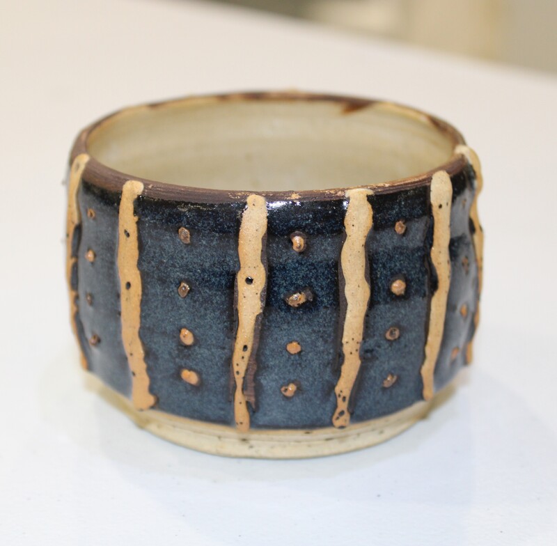 A handcrafted ceramic cup with slate-blue glaze, beige vertical lines, and dot patterns.
