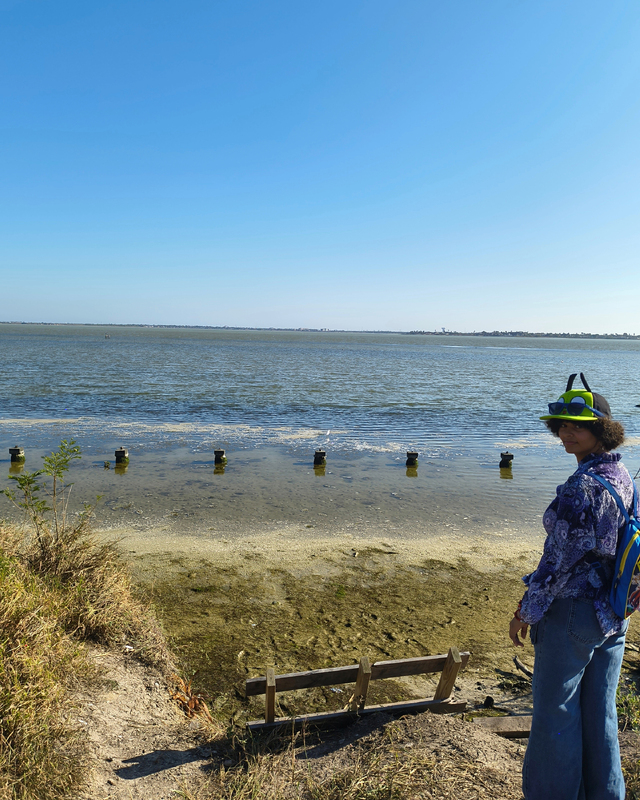 A woman in a colorful hat and shirt stands by a calm body of water with wooden posts visible in the foreground and a clear blue sky above.