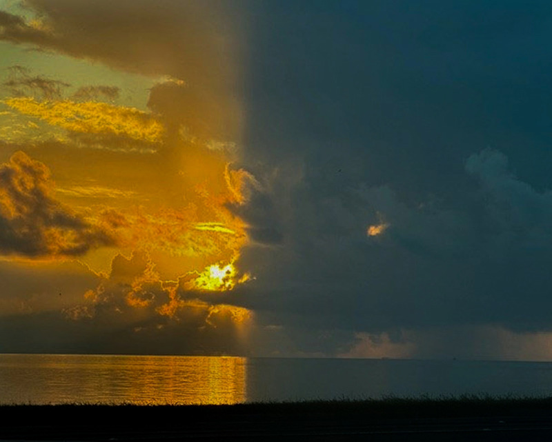 A silhouette of grassy shoreline in the foreground leads to a calm body of water. The rest of the frame is filled with a clouded sky with dramatic shadows and reflections. The clouds on the right are heavier and shade the sun's reflection, bisecting the image with sunlight in the clouds and water's surface on the left and grey shadow on the right.