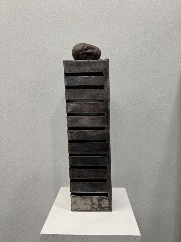 Artist: JinSun Kim. 24 by 7-inch sculpture of metal and clay. An upright tower of metal with nine slits on the left side of the piece. A sculpted head rests on top of the tower.