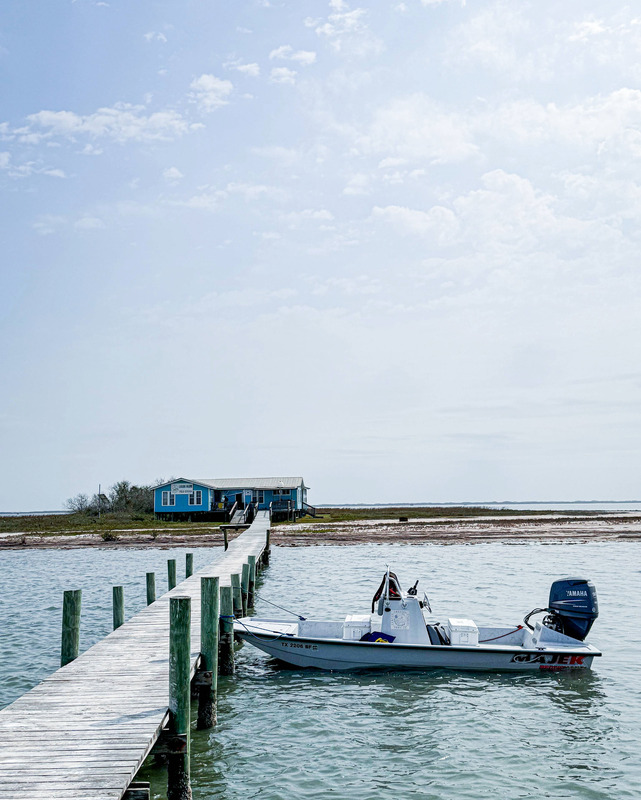 A boat with an outboard motor is tied to a wooden pier in a body of water. The pier leads to a distant shore in the background, leading away from the viewer. On the shore is a blue and white building with shrubbery and low grass. A blue sky with light clouds is visible in the background.