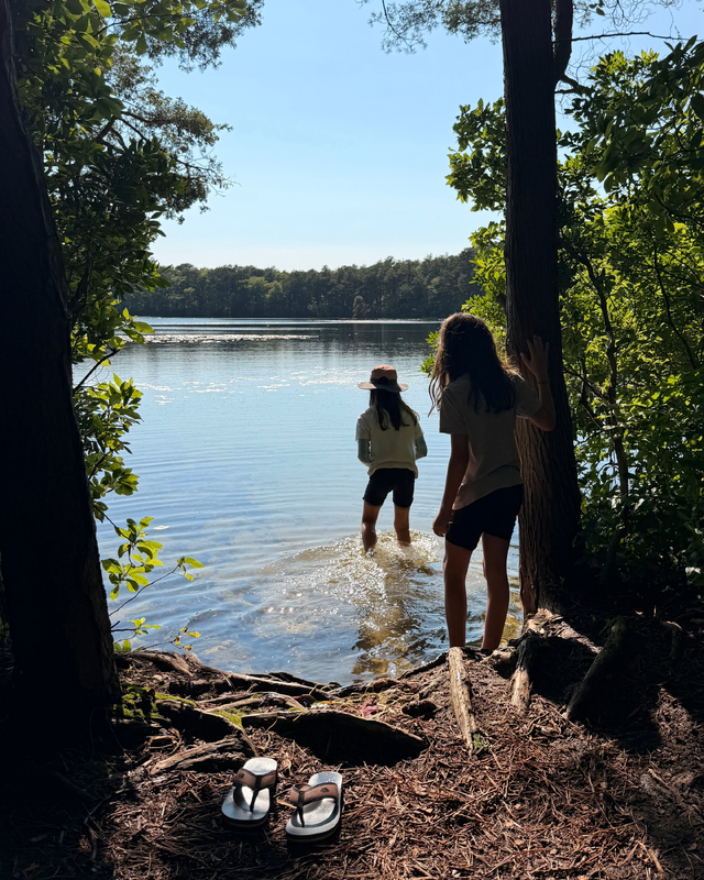 Two individuals by a tranquil lake, one wading in the water and the other standing on the shore, surrounded by greenery and a pair of flip-flops on the ground.