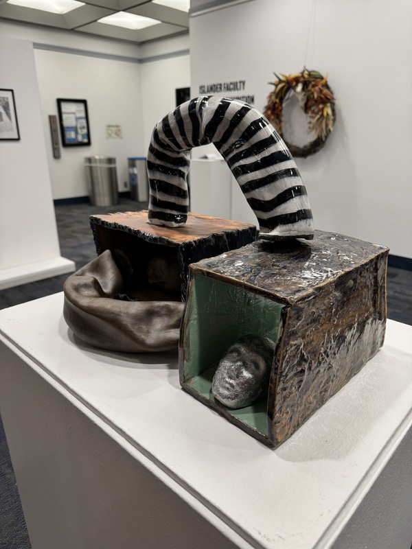 Artist: JinSun Kim. 13 by 18-inch clay work with multiple pieces. Open side rectangular sculptures sit side by side. Curved tubes of clay connect the pieces from the side and the top. A small sculpted head rests inside each box. The pieces are glazed in a variety of colors and patterns. 
