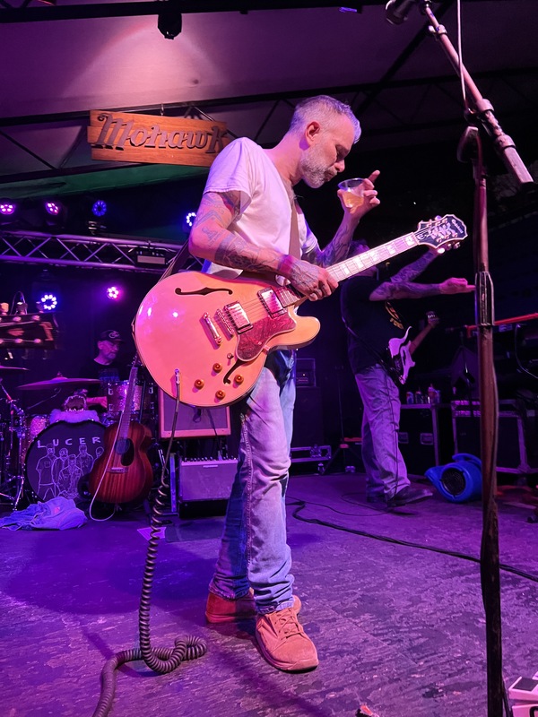 Image of a man with a guitar, taking a drink from a clear cup. The man is with other musicians on a stage at night time.