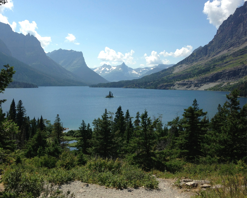 A tranquil lake surrounded by mountains, with a small island and evergreen trees in the foreground under a blue sky with white clouds.