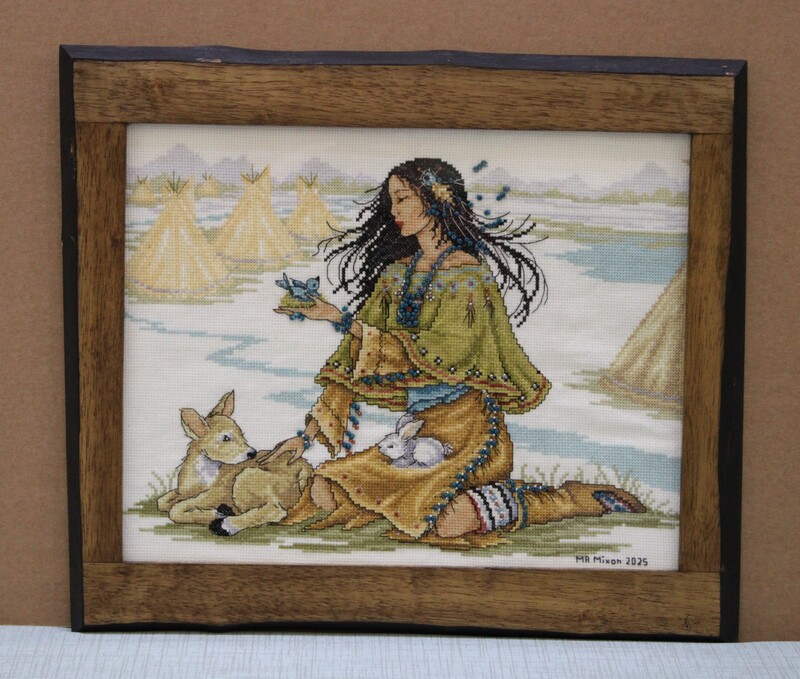 Cross-stitch art of a woman with animals in a natural setting, framed in wood.