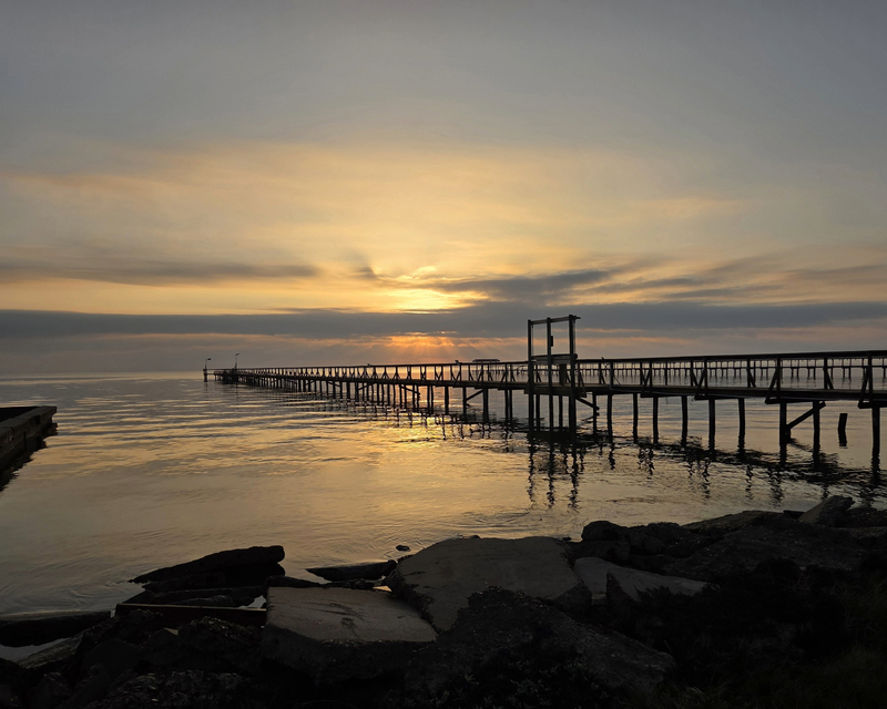 A wooden pier extends into calm water at sunset, with rocky shorelines in the foreground and a colorful sky filled with soft hues of orange and gray.