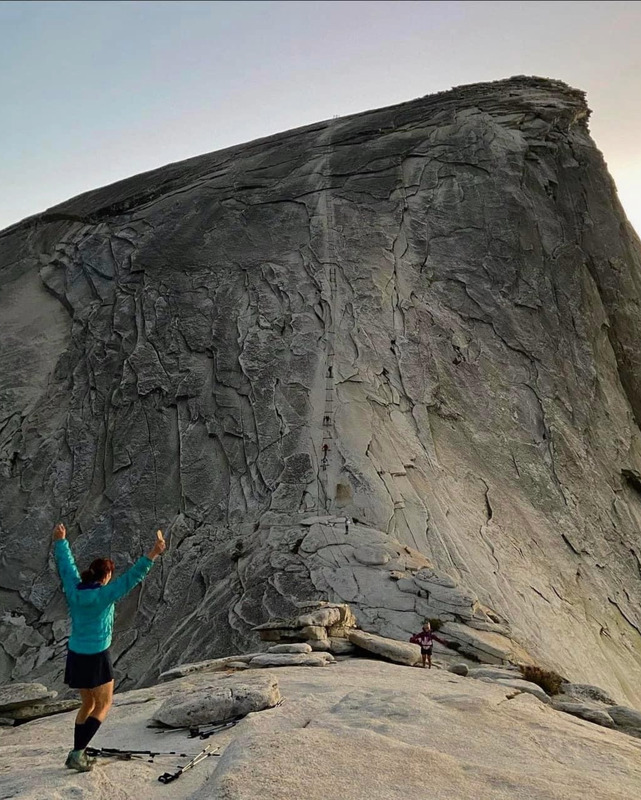 A figure stands with arms upraised facing a large rock formation. The figure is in a long sleeved shirt, black shorts, long black socks and hiking shoes, facing away from the viewer. Hiking equipment lies on the rock at the figure's feet. The trail up the rock formation has a ladder like appearance and figures can be seen on the path from a great distance. 