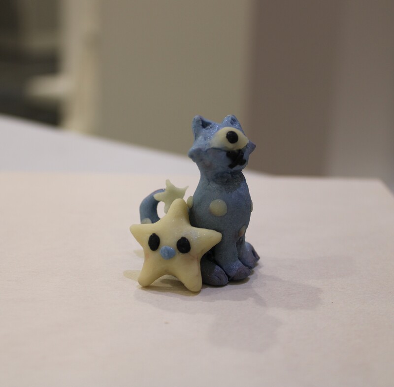 A blue cat-like sculpture sits next to a yellow star-shaped figure on a light surface.
